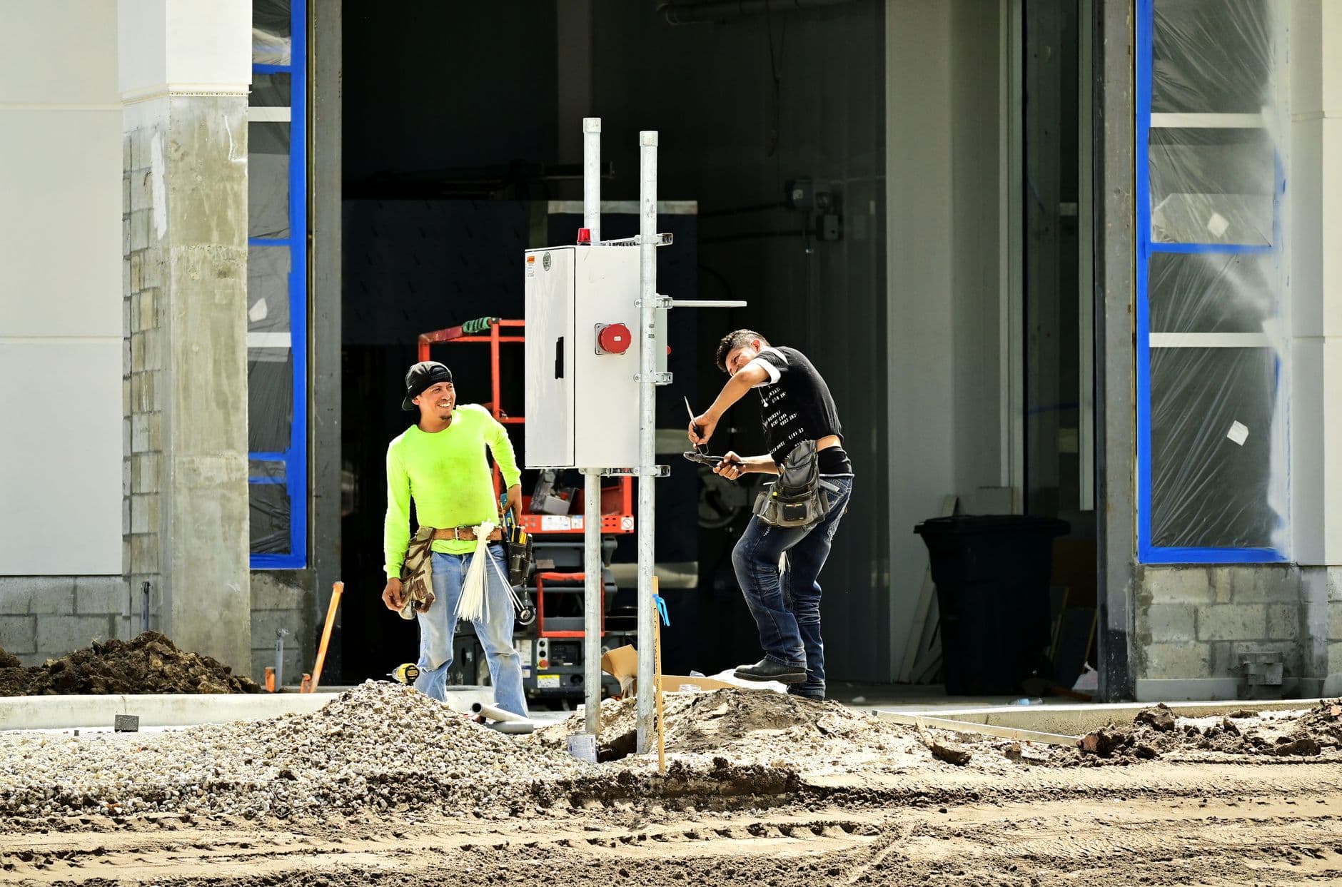 Two construction workers in hard hats installing an electrical panel at a job site